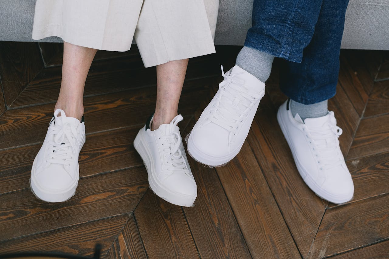 Two people wearing casual white sneakers, seated together on a wooden floor indoors.